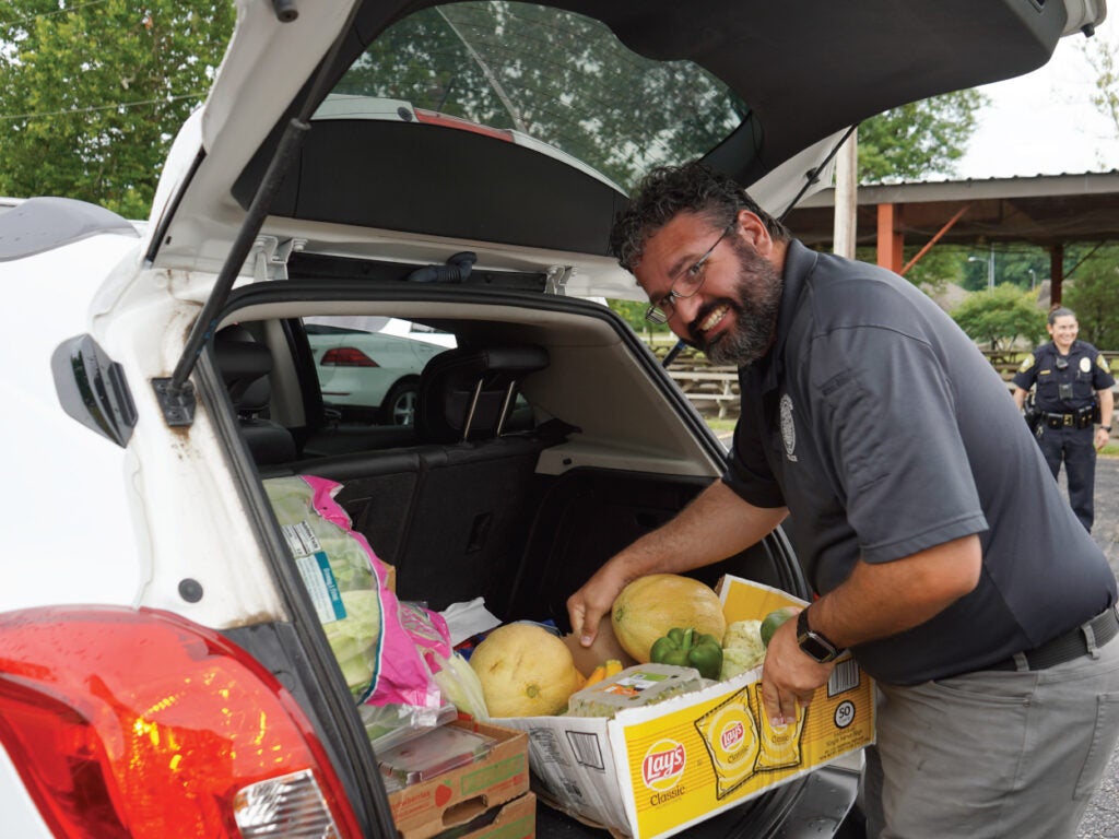 A man in glasses smiles while unloading produce from the trunk of a white car. A police officer stands behind him.